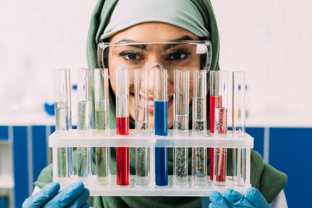smiling female muslim scientist holding test tubes with red and blue liquid while looking at camera - Fotó, kép