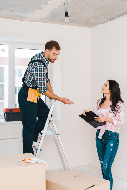 cheerful woman holding clipboard and looking at handyman standing on ladder - Φωτογραφία, εικόνα