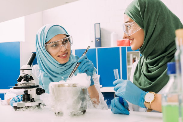Lächelnde muslimische Wissenschaftlerinnen experimentieren im Chemielabor mit Mikroskop und Trockeneis - Foto, Bild