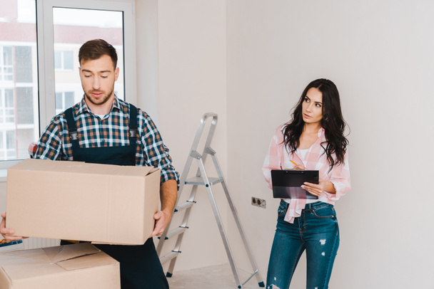 attractive woman holding clipboard and looking at handsome handyman with box in hands - Foto, Bild