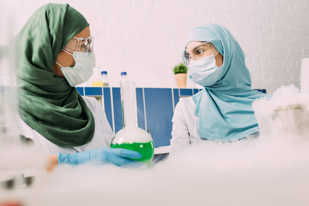 θηλυκό μουσουλμάνων επιστημόνων με φιάλη βλέπουν άλλο πειραματιμένος με ξηρό πάγο σε χημικό εργαστήριο - Φωτογραφία, εικόνα