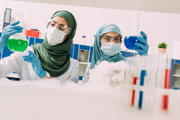 化学実験室でドライアイスを試しながらフラスコを持って医療マスクで女性イスラム教徒の科学者 - 写真・画像