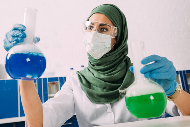 female muslim scientist in medical mask holding flasks during experiment in chemical laboratory - Foto, Imagen