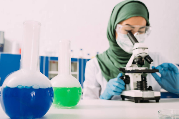 γυναίκα μουσουλμάνος επιστήμονας κάθεται στο τραπέζι με φιάλες και χρησιμοποιώντας μικροσκόπιο κατά τη διάρκεια του πειράματος σε χημικό εργαστήριο - Φωτογραφία, εικόνα