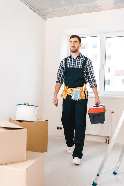 handsome handyman holding toolbox and walking near boxes in room - Foto, Bild