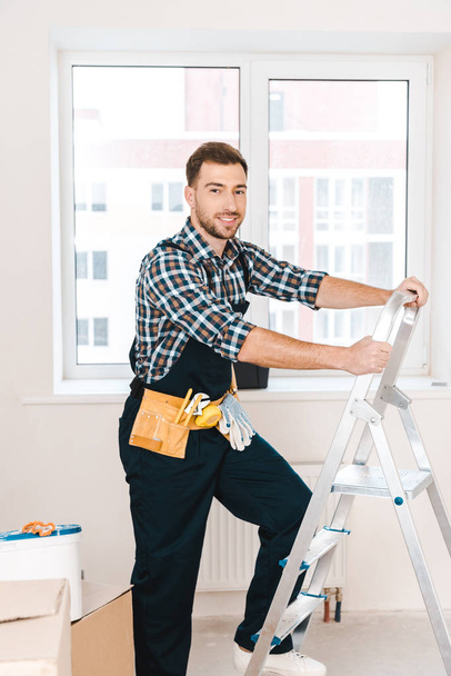 cheerful handyman standing near ladder and smiling in room - Photo, Image