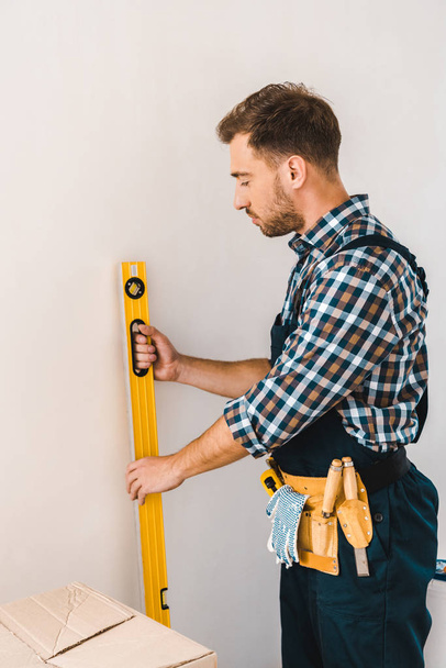 handsome handyman holding measuring level near wall - Foto, Imagem