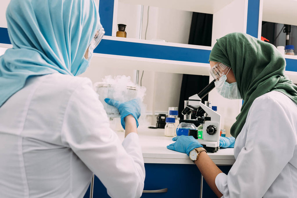θηλυκό μουσουλμάνων επιστημόνων πειραματίζεται με τον ξηρό πάγο και χρησιμοποιώντας μικροσκόπιο σε χημικό εργαστήριο - Φωτογραφία, εικόνα