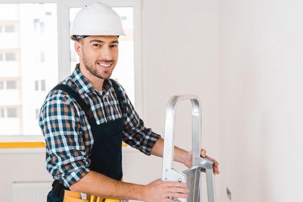 cheerful handyman in helmet smiling while holding ladder  - Photo, Image
