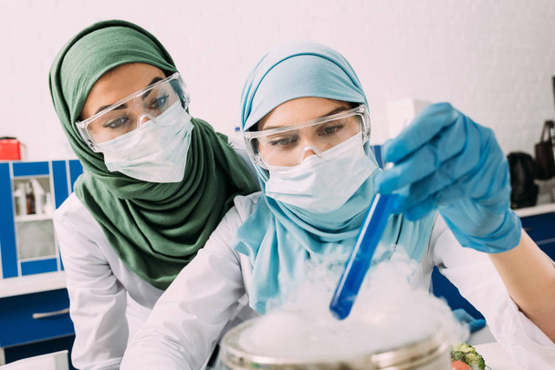 female muslim scientists in goggles holding reagent over dry ice during experiment in chemical lab - Photo, Image