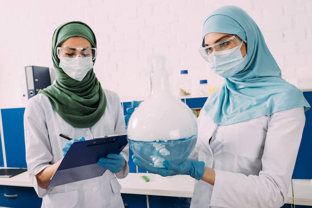 científicos musulmanes femeninos concentrados en máscaras médicas que sostienen el portapapeles y el líquido de crecimiento del frasco en el laboratorio químico
 - Foto, Imagen