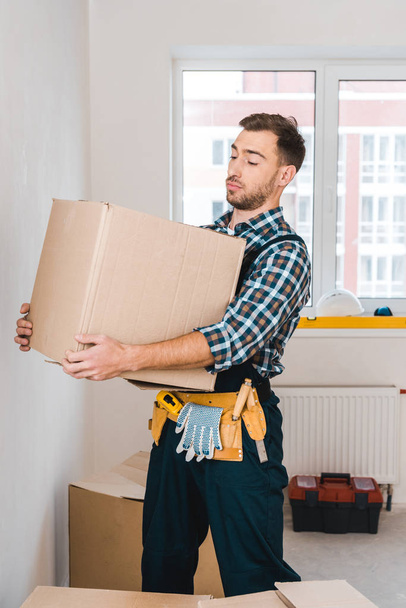 handsome handyman holding box while standing in room - Photo, Image