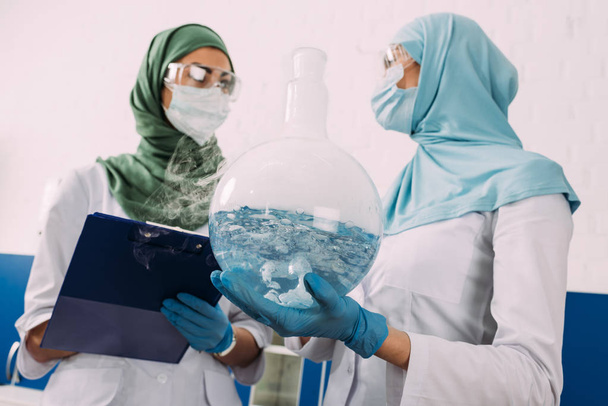 scienziati musulmani femminili che tengono appunti e liquido di crescita della fiaschetta in laboratorio chimico
 - Foto, immagini