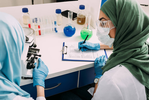 θηλυκό μουσουλμάνοι επιστήμονες χρησιμοποιώντας μικροσκόπιο και γράφοντας στο πρόχειρο κατά τη διάρκεια του πειράματος σε χημικό εργαστήριο - Φωτογραφία, εικόνα