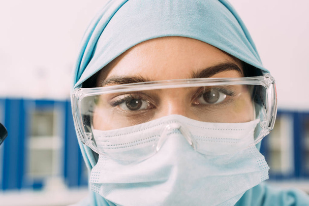 close up of female muslim scientist in medical mask, goggles and hijab looking at camera  - Zdjęcie, obraz