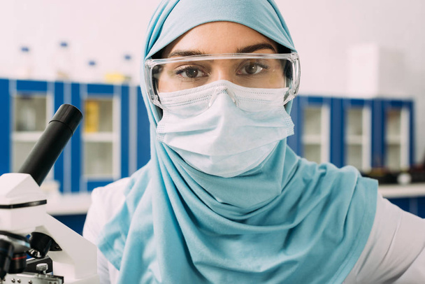 γυναίκα μουσουλμάνος επιστήμονας στην ιατρική μάσκα, προστατευτικά δίοπτρα και χιτζάμπ βλέπουν φωτογραφική μηχανή στο εργαστήριο - Φωτογραφία, εικόνα