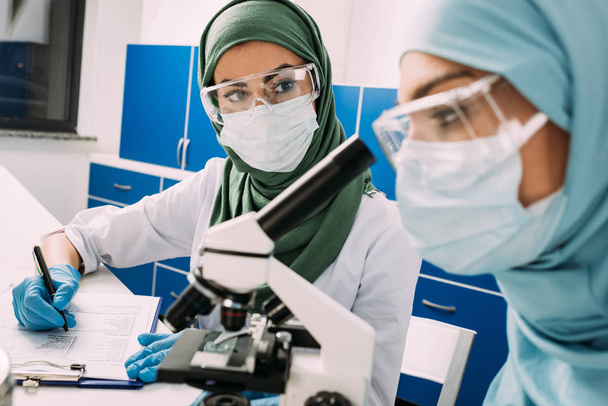 mulheres cientistas muçulmanas usando microscópio e escrita em prancheta durante experimento em laboratório químico
 - Foto, Imagem