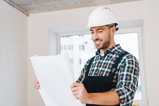 cheerful handyman smiling while looking at blueprint - Foto, afbeelding