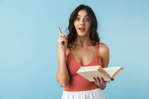 Lovely girl wearing summer clothes standing isolated over blue background, holding a book - Foto, afbeelding