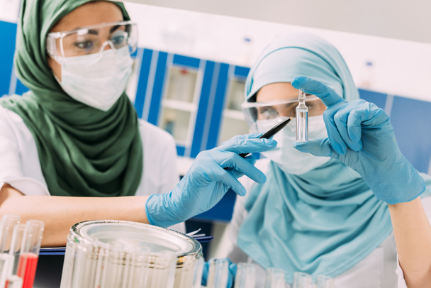 vrouwelijke islamitische wetenschappers in de medische maskers holding glazen ampul tijdens experimenteren in chemisch laboratorium - Foto, afbeelding