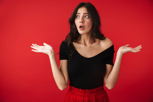 Photo of a beautiful confused shocked young woman standing isolated over red wall background. - Foto, immagini