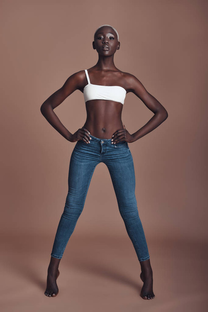 Full length of attractive young African woman looking at camera and keeping hands on hips while standing against brown background - Фото, изображение