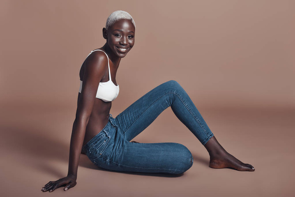 happy smiling African woman sitting against brown background and wearing top with denim jeans  - Fotoğraf, Görsel