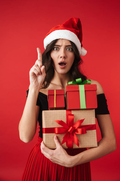 Excited young woman wearing Christmas hat standing isolated over red background, holding stack of gift boxes - Foto, afbeelding