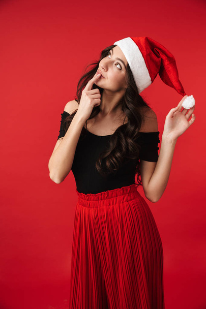 Захоплена молода жінка в різдвяному капелюсі стоїть ізольовано на червоному тлі, позує
 - Фото, зображення