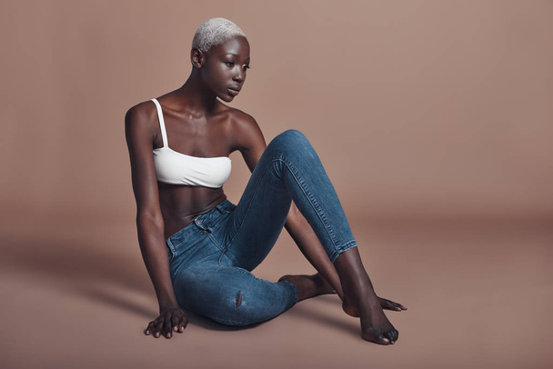 Attractive barefoot African woman sitting against brown background and wearing top with denim jeans  - Фото, изображение