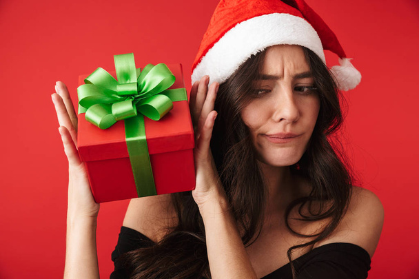 Upset young woman wearing Santa hat holding gift box isolated over red background - Fotoğraf, Görsel