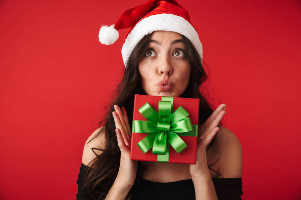 Excited young woman wearing Santa hat holding gift box isolated over red background - Valokuva, kuva