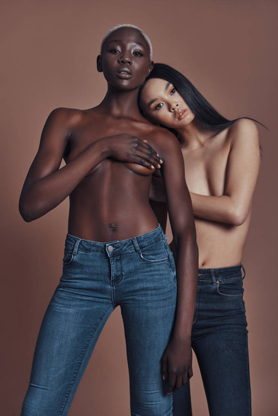 two shirtless women in denim jeans posing against brown background - Foto, afbeelding