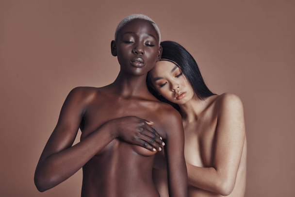 two shirtless women posing against brown background and covering breast with closed eyes  - Foto, Imagem