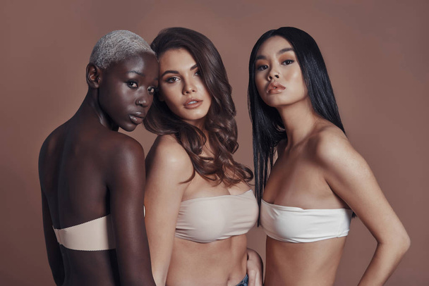 three women wearing bras and posing against brown background and looking at camera - Photo, image