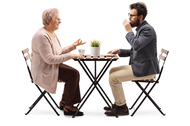 Full length profile shot of a senior woman sitting at a table with coffee and talking to a young bearded man isolated on white background - Valokuva, kuva