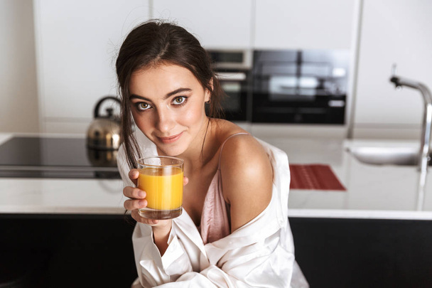 Photo of caucasian woman 20s wearing silk leisure clothing drinking juice in kitchen - Photo, Image