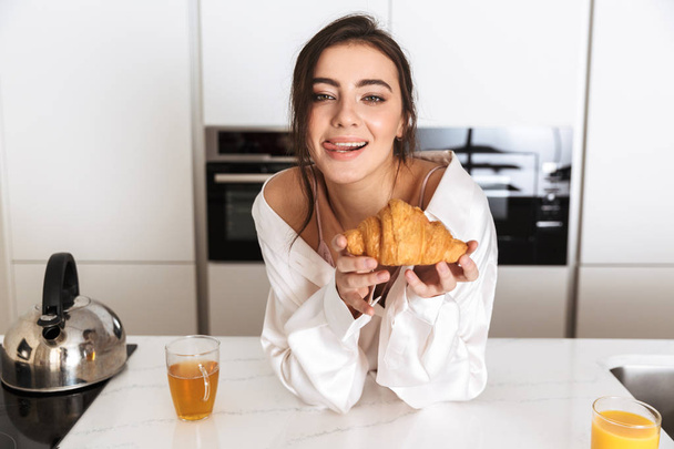 Photo of caucasian woman 20s wearing silk leisure clothing having breakfast in kitchen - Photo, image