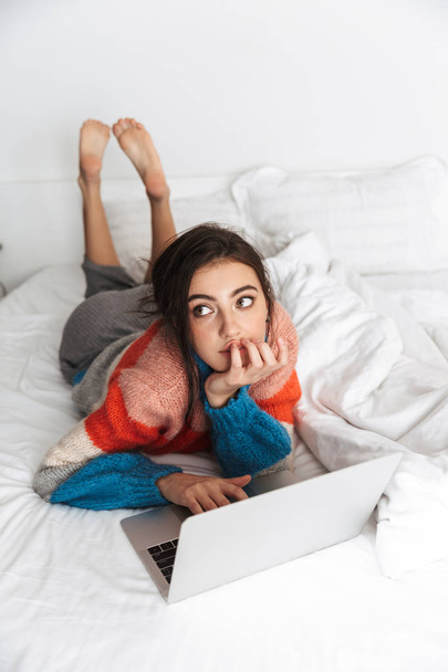 Retrato de mujer joven con suéter acostado en el sofá en el apartamento y el uso de la computadora portátil
 - Foto, Imagen