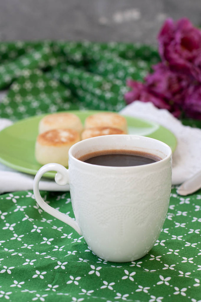 Black coffee in a white sophisticated cup, delicious dietary cheesecakes from home-made farmer cheese for breakfast, a bouquet of purple tulips on a green tablecloth. - Photo, Image