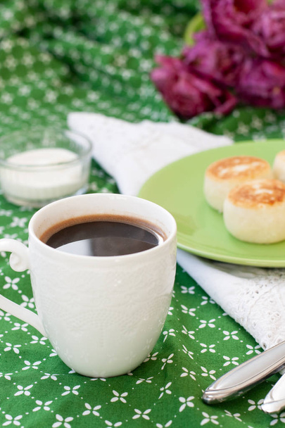 Black coffee in a white sophisticated cup, delicious dietary cheesecakes from home-made farmer cheese for breakfast, a bouquet of purple tulips on a green tablecloth. - Photo, Image
