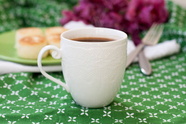 schwarzer Kaffee in einer weißen, raffinierten Tasse, köstliche diätetische Käsekuchen aus hausgemachtem Bauernkäse zum Frühstück, ein Strauß lila Tulpen auf einer grünen Tischdecke. - Foto, Bild