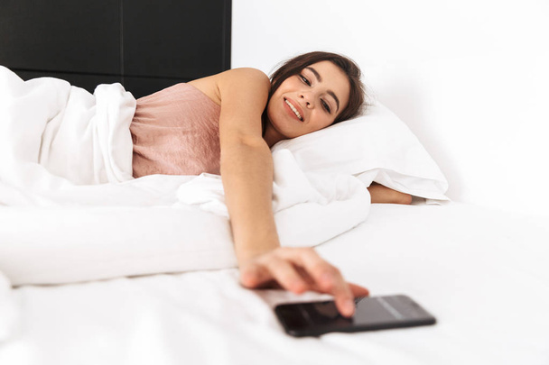Photo of pretty woman 20s with dark hair holding smartphone while lying in bed on white pillow after sleep or nap - Foto, Bild