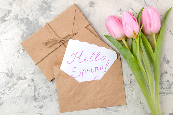 ein Strauß schöner rosafarbener Tulpenblüten auf hellem Betongrund. Frühling. Urlaub. Text Hallo Frühling auf Papier in einem Umschlag. Ansicht von oben - Foto, Bild