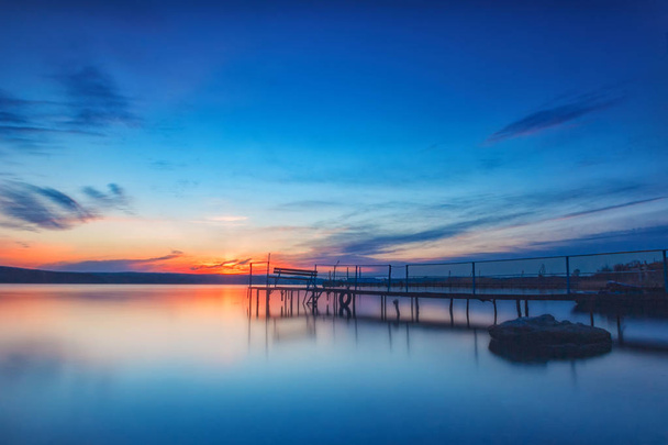 Geweldige lange blootstelling zonsondergang aan het meer met een bankje op de houten pier. - Foto, afbeelding