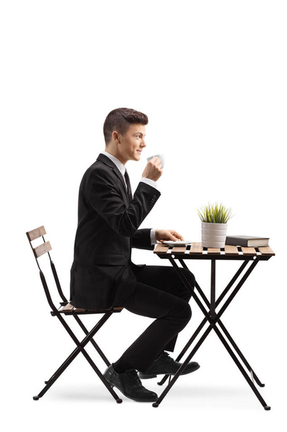 Full length profile shot of a young man in a suit sitting and drinking coffee isolated on white background - Φωτογραφία, εικόνα