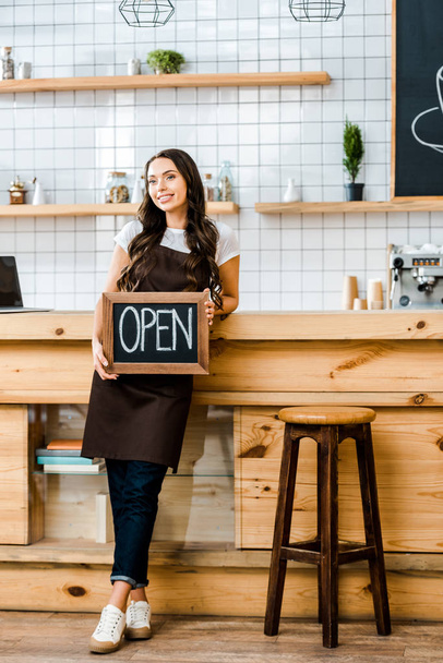 beautiful cashier standing near wooden bar counter and holding chalkboard with open lettering in coffee house - Foto, immagini