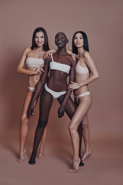 Full length of three attractive mixed race women looking at camera and smiling while standing against brown background - Foto, Bild