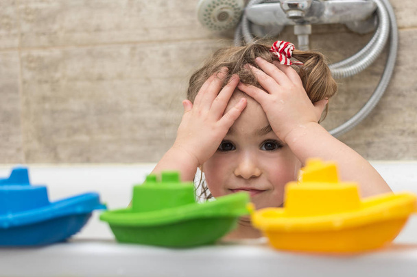 Happy babymeisje zitten in badkuip spelen met kleurrijke boten speelgoed in de badkamer. Glimlachend kind in badkamer op bruine achtergrond. Baby wassen en Baden. Hygiëne en gezondheidszorg.  - Foto, afbeelding