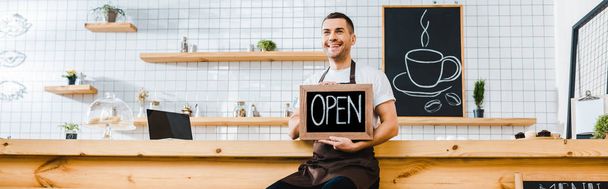 handsome cashier in brown apron sitting on chair near wooden bar counter and holding chalkboard with open lettering in coffee house - Photo, Image